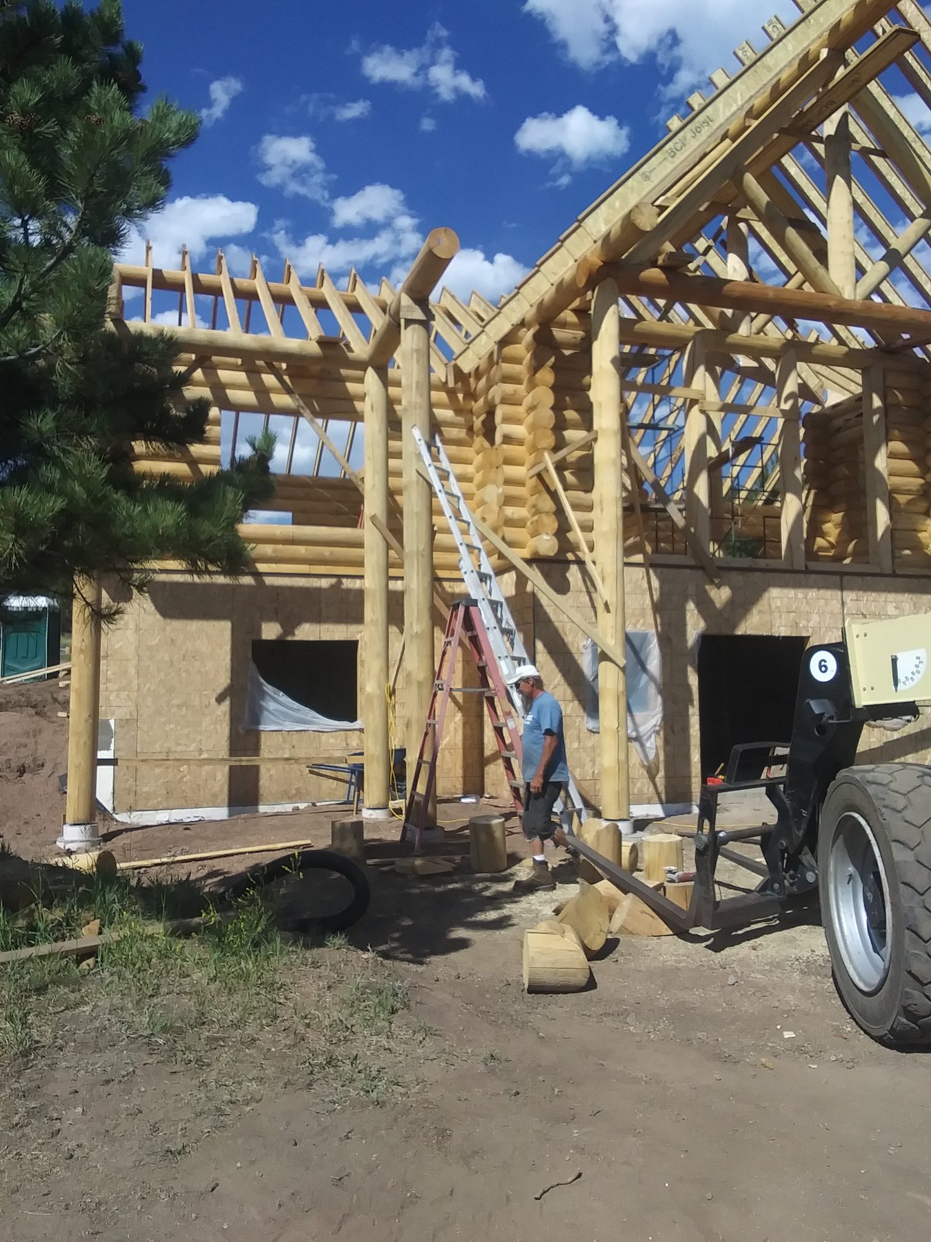 Timber trusses being raised over the log walls, with a telehandler holding the lift.